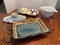 Overview of ceramic servingware pieces on wooden table with neutral white backdrop including vegetable and dip tray on metal stand, divided fruit serving platter, and white grape bowl.
