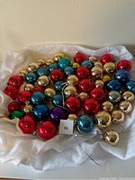 Overhead view of approximately 60 vintage glass Christmas ball ornaments in shiny metallic red, gold, blue, green and purple, arranged on white fabric, showing varying sizes and minor wear.