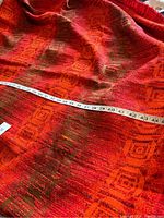 Photo showing the fabric with a measuring tape along the length to indicate dimensions of approximately 76 inches. The fabric has an orange and brown geometric square pattern with textured weave.