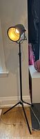 Full view of black floor lamp standing on hardwood floor with light turned on, showing tripod base and cord extending from base.