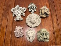 All seven wall decor pieces arranged on a wooden table showing varied gargoyle faces, the cherub medallion, basket, and lion face