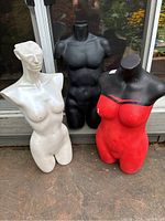 Group photo showing all three mannequin torsos standing outdoors on a concrete surface. White female torso, black hollow male torso, and red female torso arranged together.