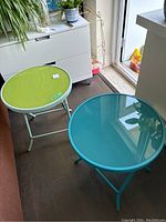 Photo showing both tables side by side indoors near a cabinet, exhibiting bluey turquoise and green glass tops with metal frames, folded and set for display.