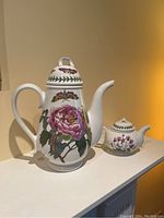 View of coffee pot with pink peony and butterfly design along with smaller matching tea-for-one teapot on white surface