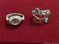 Two sterling silver rings shown on red surface; left ring has a large oval faceted light brown stone, right ring has colorful small stones with copper accents