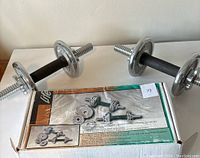 Two assembled dumbbells with chrome plates and textured black handles alongside original product box showing weight plate and collar details.