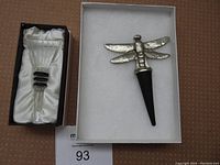 Metal dragonfly wine stopper with black rubber stopper in white case on right; glass stopper with rubber rings in black box on left.