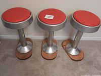 Three antique ice cream stools side by side showing red cushioned seats, silver metal trim and wooden bases
