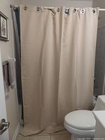 Beige/cream shower curtain hanging on a shower rod inside a bathroom, showing front view with metal grommets at top and curtain hanging down close to floor.