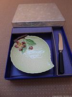 Photo of original box with cream lid opened showing ceramic leaf-shaped dish with flower and fruit decoration and a metal spreader with black handle resting inside molded compartments.