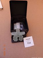 Top view of the light green glass Inukshuk figurine resting inside a black lined box including accompanying description card and MaxSold lot number 104.
