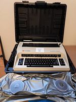 Olympia electronic compact typewriter inside black plastic hard case with attached cord. Seen full front and top view showing keyboard and function keys.