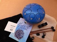 Blue metal tongue drum with accessories including drumsticks, finger pads, drumstick holder, musical score booklet, and two pouches on a wood table.