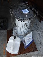 Front view of the white kerosene heater with metal cage, showing the attached handle and vents. The heater is placed on a wooden board. Manual and white plastic jug are in front of the heater.