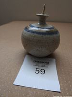 Front and side view of pottery oil lamp showing glazed bands on round body, wick holder on top, placed on beige surface with label.