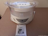 Full view of Rival electric food steamer showing three clear plastic baskets stacked and lid on top, placed on a surface. Instruction booklet is in front of steamer.
