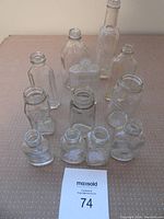 Front view of the 12 clear glass bottles on a fabric surface showing different heights and shapes, open necks visible