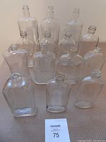 Top view showing a collection of 14 clear glass bottles with various surface patterns and plain glass, arranged in rows on a surface with a MaxSold lot identifier visible.