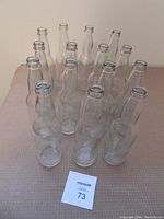 Front view of 15 clear glass bottles arranged in a tight cluster on a beige surface with a MaxSold lot number paper in front.