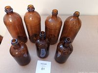 Front view showing 7 brown glass bottles of differing heights arranged upright showing embossed lettering.