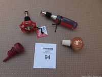 Set of four wine stoppers shown on beige surface with MaxSold auction sign. Includes redneck valve handle, screwdriver-shaped stopper, red plastic stopper, and wooden deer silhouette stopper.
