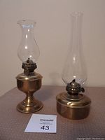 Two vintage brass and glass oil lamps with wicks, one smaller with scalloped glass chimney and one larger with tall cylindrical glass chimney, photographed against a plain background with a MaxSold lot card.
