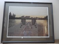 Framed print showing two wooden Adirondack chairs on a dock with a small table holding wine glasses, overlooking a lake and surrounded by trees at sunset. The print is matted and framed under glass.