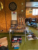 Wide shot showing dartboard on the wall above scoreboard, three wooden tennis rackets leaning against the wall, backgammon set open on folding table with pieces visible, bocce ball set in box below table, and additional game accessories and board games nearby.