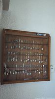 Front view of wooden oak display case mounted on wall, containing approximately 70 collectible spoons arranged in rows with various decorative handles.