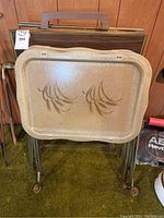 Photo of stacked metal TV tables with a beige painted metal tray top featuring a brown decorative pattern and folding chrome legs with some legs having castor wheels.