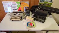 Two typewriters on a white table surrounded by boxes of photo and printer paper, lens cleaning kit, and small slide viewer.