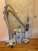Photo of Electrolux vacuum cleaner, steam mop, and vacuum attachments on wooden floor in front of a wall.
