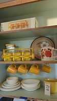 Overall view of shelves with yellow Corning Ware mugs hanging, stacked plates and bowls below with a floral design teapot and revolving spice rack on upper shelf.