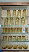 Full view of all vintage amber glassware on glass shelves, including 10 tall tumblers, 4 short tumblers, and 4 small bowls at bottom shelf.