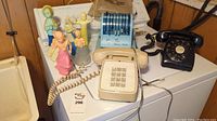 Wide shot showing all items on a white surface against wood paneling: three vintage telephones, Paymaster Ribbon Writer cheque writer, and six Avon perfume bottles in different shapes and pastel colors.