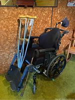 Photo showing black wheelchair with two wooden crutches and a metal cane placed in front, angled view showing padded seat and armrests.