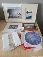 Photo showing four framed nature art pieces on a table alongside a blue and white decorative plate partially wrapped in bubble wrap with some packaging material visible.