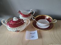 Photo showing creamer jug, sugar bowl with lid, and soup bowl on its plate all in red, white, and gold finish with floral design.