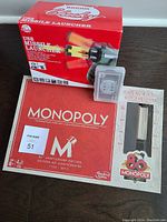 Monopoly 80th Anniversary Edition game box, deck of cards in plastic case, and boxed USB missile launcher placed on plain surface.