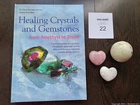 Photo showing the book titled 'Healing Crystals and Gemstones From Amethyst to Zircon' with an image of crystals on the cover, laid on a wooden surface alongside a lot number card.