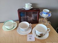Overview of 7 cups with saucers in various floral and geometric designs, plus wooden tea box