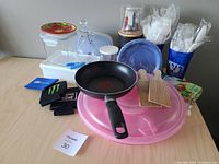 Overview of mixed kitchen items including frying pan, plastic cutlery in blue containers, paper plates, plastic and glass containers and wooden spatulas