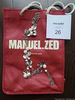 Manuel.Zed necklace partially folded on original red gift bag with white brand print and rope handles visible