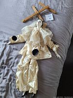 White-faced marionette puppet laid flat with wooden control bar and strings visible, showing cream satin outfit with ruffled collar and black circular accents.