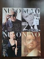 Four issues of Nuvo Designer Magazine arranged on a dark wood floor, showing front covers with celebrity photos: Michael Kors, Chris Botti, Naomi Watts, and Elijah Wood.