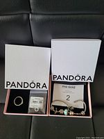 Two open Pandora boxes showing a silver ring and unopened charm in one box, and a silver bracelet with six assorted charms in another.