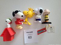 Top view showing all 5 Peanuts figurines in a line on light surface including ceramic Charlie Brown with tags, Snoopy in Joe Cool shirt, Woodstock, classic Snoopy, and Snoopy on doghouse.