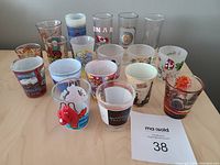 Eighteen assorted shot glasses arranged on a table, showing various travel and souvenir designs from around the world including Canada, animals, city scenes, and logos.