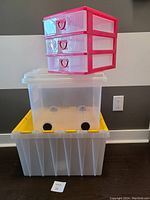 Stacked storage containers: top is red 3-drawer unit, middle is generic plastic container with lid and wheels, bottom is large translucent plastic container with yellow lid