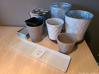 Overview of pots and trays including white, beige, and gray ceramic pots with textured surfaces; long rectangular ceramic tray and small square metallic trays visible; black plastic pots stacked inside one ceramic pot.